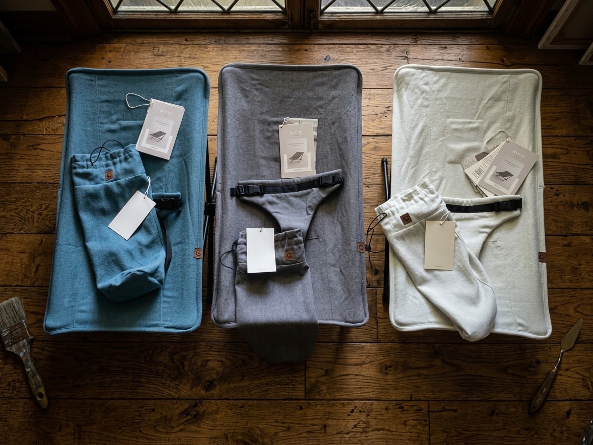 Three baby chairs laid out side by side on a wooden floor: a teal fabric seat; a gray fabric seat, and a beige fabric seat. Each has a small paper brand tag attached. Each has a matching carry case for the fabric seat and the aluminum and collapsable chair frame.